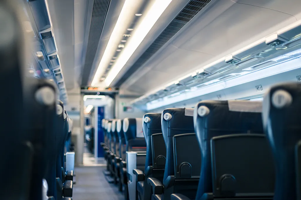 Photograph looking down the aisle of an empty, modern passenger train carriage. Rows of dark blue seats with headrest covers and folding tray tables line both sides beneath bright overhead strip lighting.