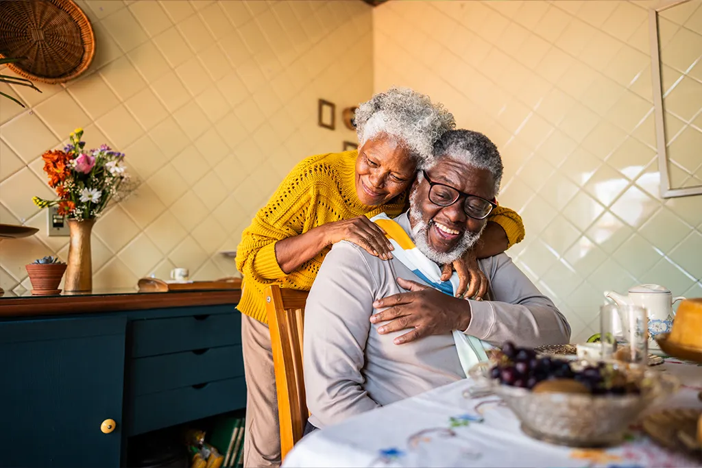 Photograph of a happy older couple sharing an affectionate moment in a warm kitchen or dining room. The woman in a yellow sweater smiles warmly as she leans over to embrace the laughing man seated at the breakfast table.