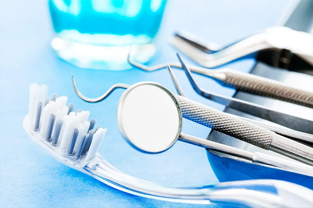 Close-up photograph of dental hygiene tools on a light blue surface. A white toothbrush, a metal dental mirror, and probes rest in the foreground, with a glass of blue mouthwash blurred in the background.