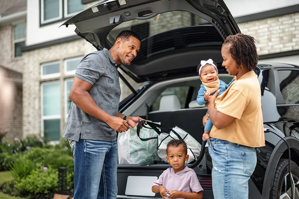 Photograph of a family gathered behind their open car trunk outside a modern suburban house. The father smiles while unloading large bags, while the mother holds a smiling baby girl alongside their young son standing in front.