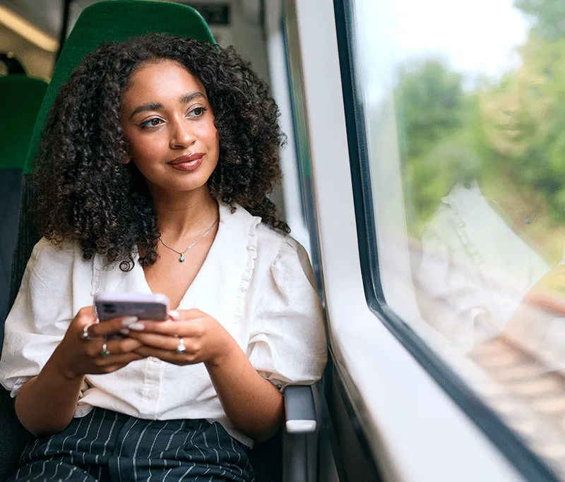Photograph of a young woman with dark curly hair sitting by the window inside a train carriage. Holding a smartphone in her hands, she smiles gently as she gazes out at the passing green landscape.