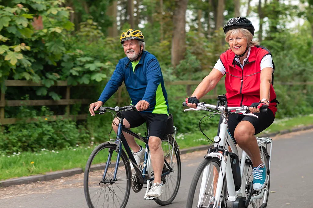 Photograph of an active senior couple smiling and riding bicycles outdoors along a paved road bordered by lush green trees. Both wear helmets and athletic clothing, with the man riding a dark bicycle and the woman riding a white e-bike.