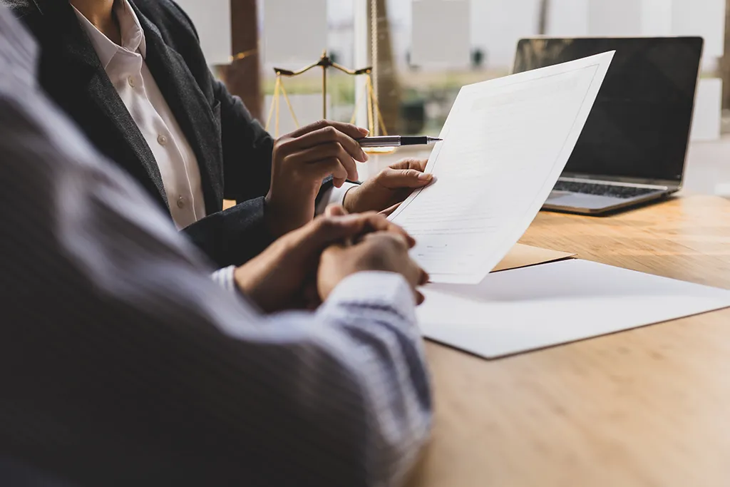 Photograph taken over the shoulder of a client reviewing legal documents with a professional across a wooden desk. A brass scale of justice and an open laptop sit in the background.