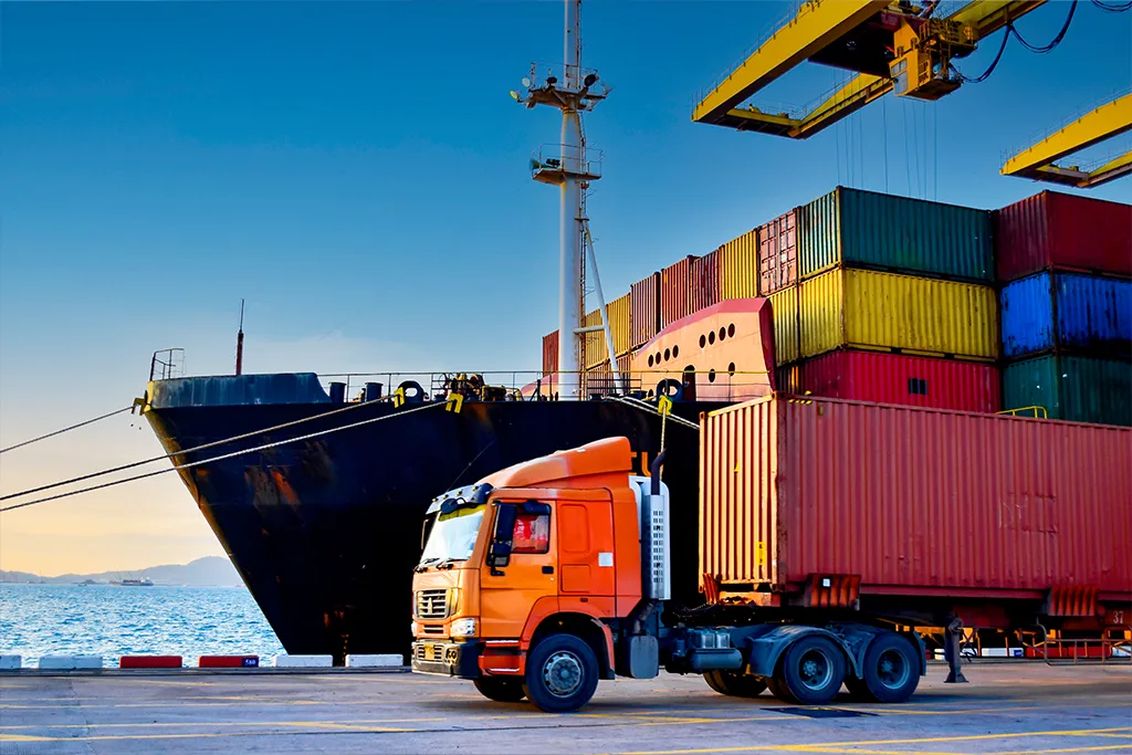 Photograph of a large orange freight semi-truck parked at a commercial shipping port, with a loaded container cargo ship docked beside it under large yellow harbor cranes.
