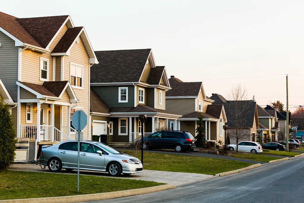 Photograph of a quiet suburban street lined with modern single-family houses with manicured front lawns. A silver sedan and a dark SUV are parked in the driveways under bright afternoon sunlight.