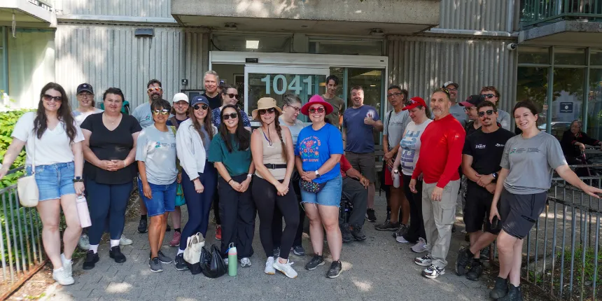 Group photograph of Engineers Canada volunteers posed outside the entrance of a Community Housing building. The group includes about two dozen people dressed in casual work attire, smiling at the camera outdoors on a sunny day.