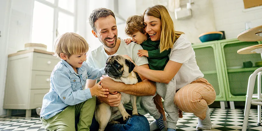 Photograph of a happy family gathered together on the tiled floor of a bright kitchen. A father, mother, and two young children surround and pet a small pug dog, smiling and laughing together.