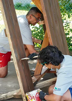 Photograph of two volunteers working outdoors to stain or paint a wooden structure. A man on his knees and a woman in the foreground wearing a light grey shirt and black gloves carefully apply wood stain with a brush.
