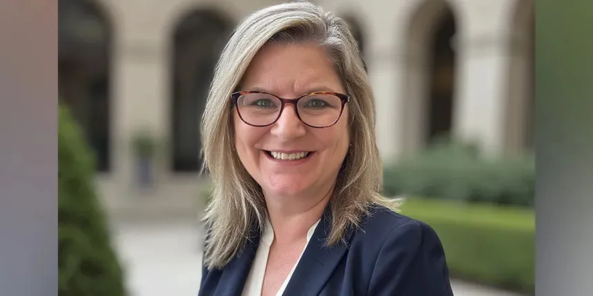 Portrait professionnel de Darlene Spracklin-Reid, présidente d’Ingénieurs du Canada, souriante en plein air. Elle porte des lunettes à monture écaille, un blazer foncé et un haut blanc, sur fond d’une cour et d’une façade de bâtiment légèrement floues.