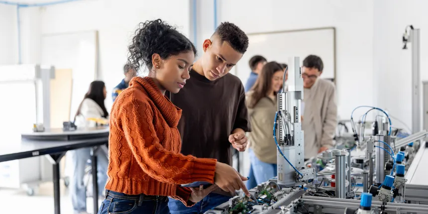Deux étudiants en ingénierie se penchent sur des composants robotiques complexes dans un laboratoire lumineux, collaborant sur un système automatisé pendant que d'autres camarades de classe travaillent en arrière-plan.